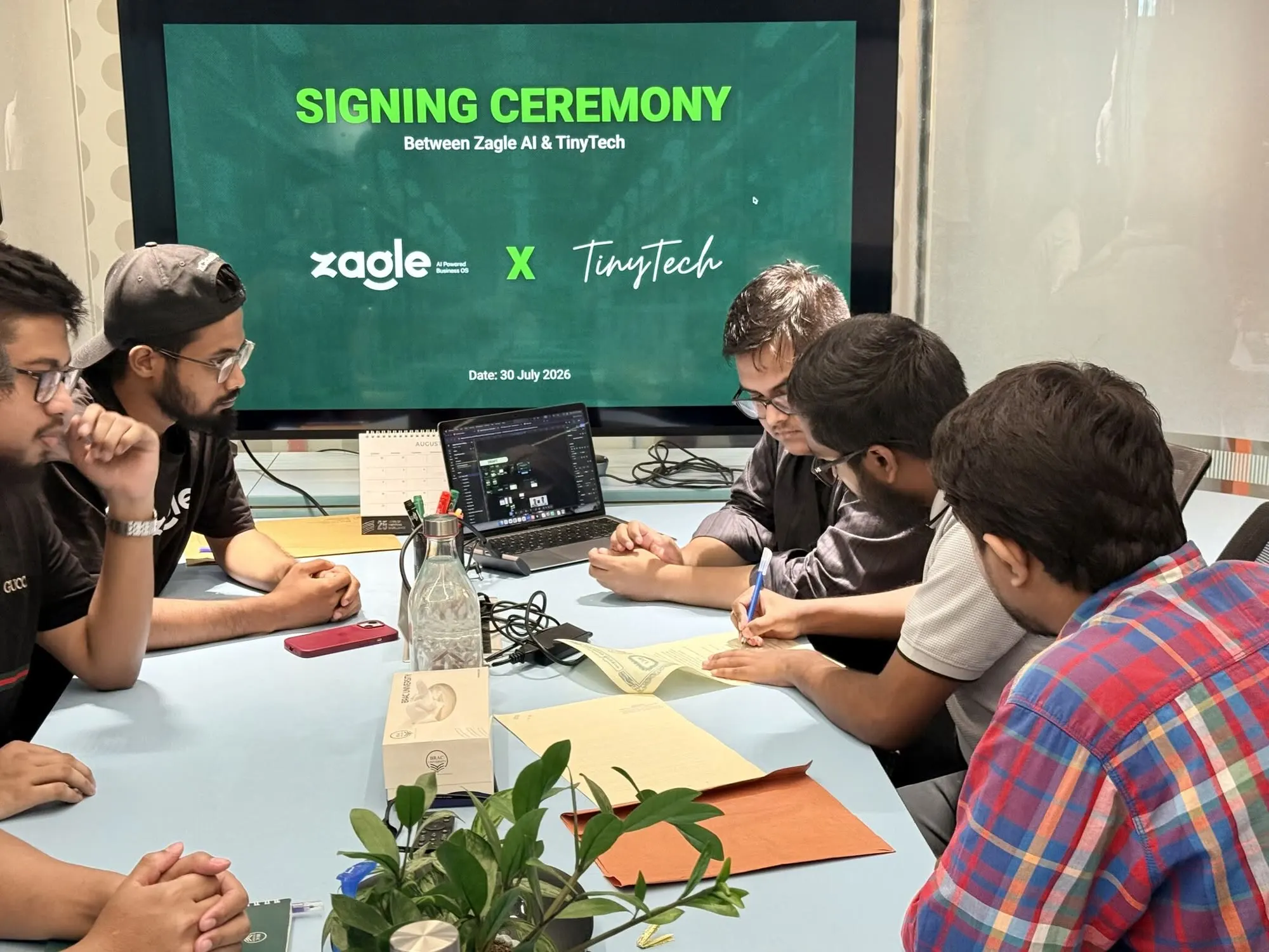 TinyTech's team signing the agreement at the table while the Zagle team looks on.
