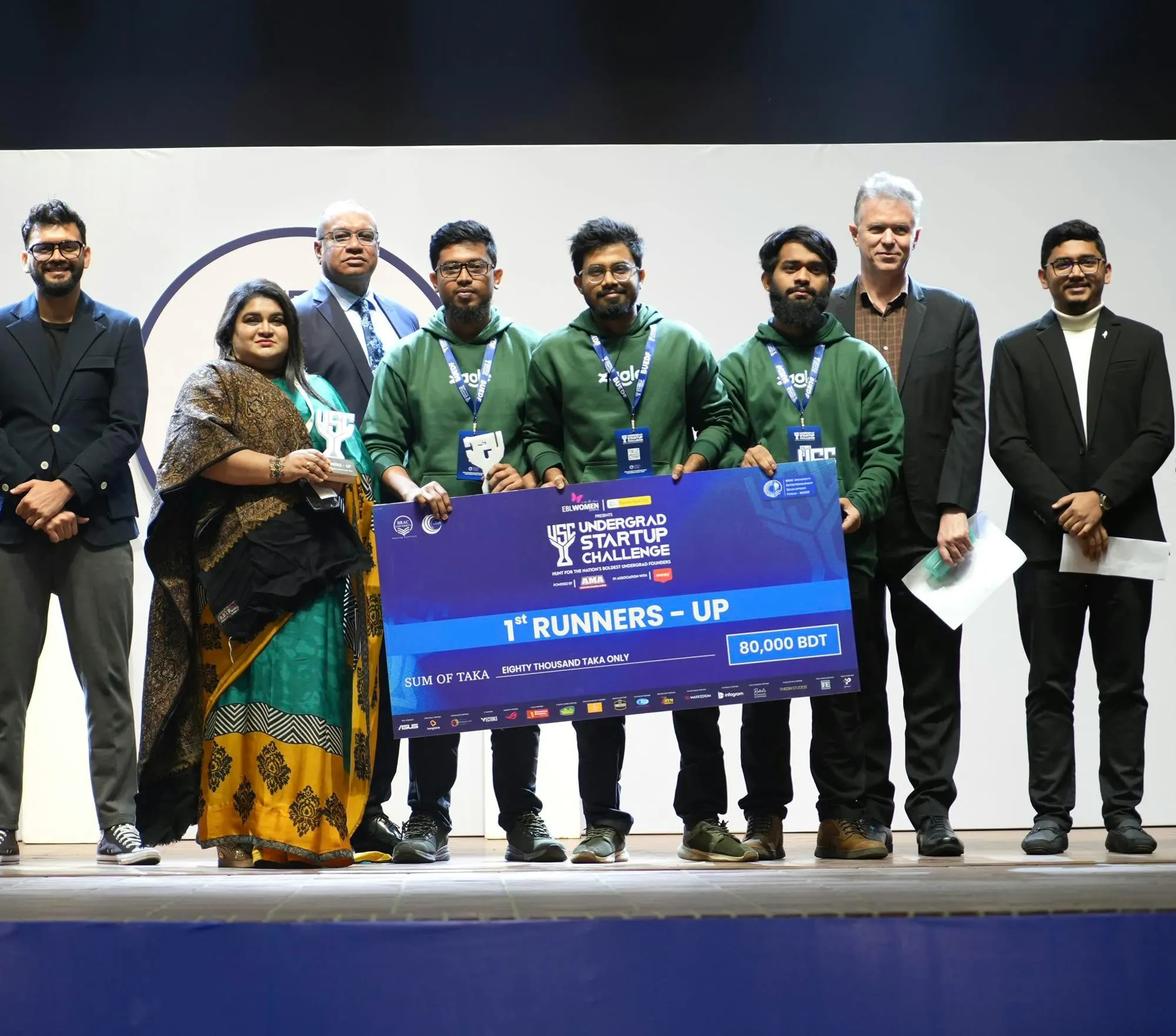 The Zagle founders in green Zagle hoodies on stage with the organisers and judges, holding a cheque for 80,000 taka for first runners-up at the EBL Undergrad Startup Challenge.