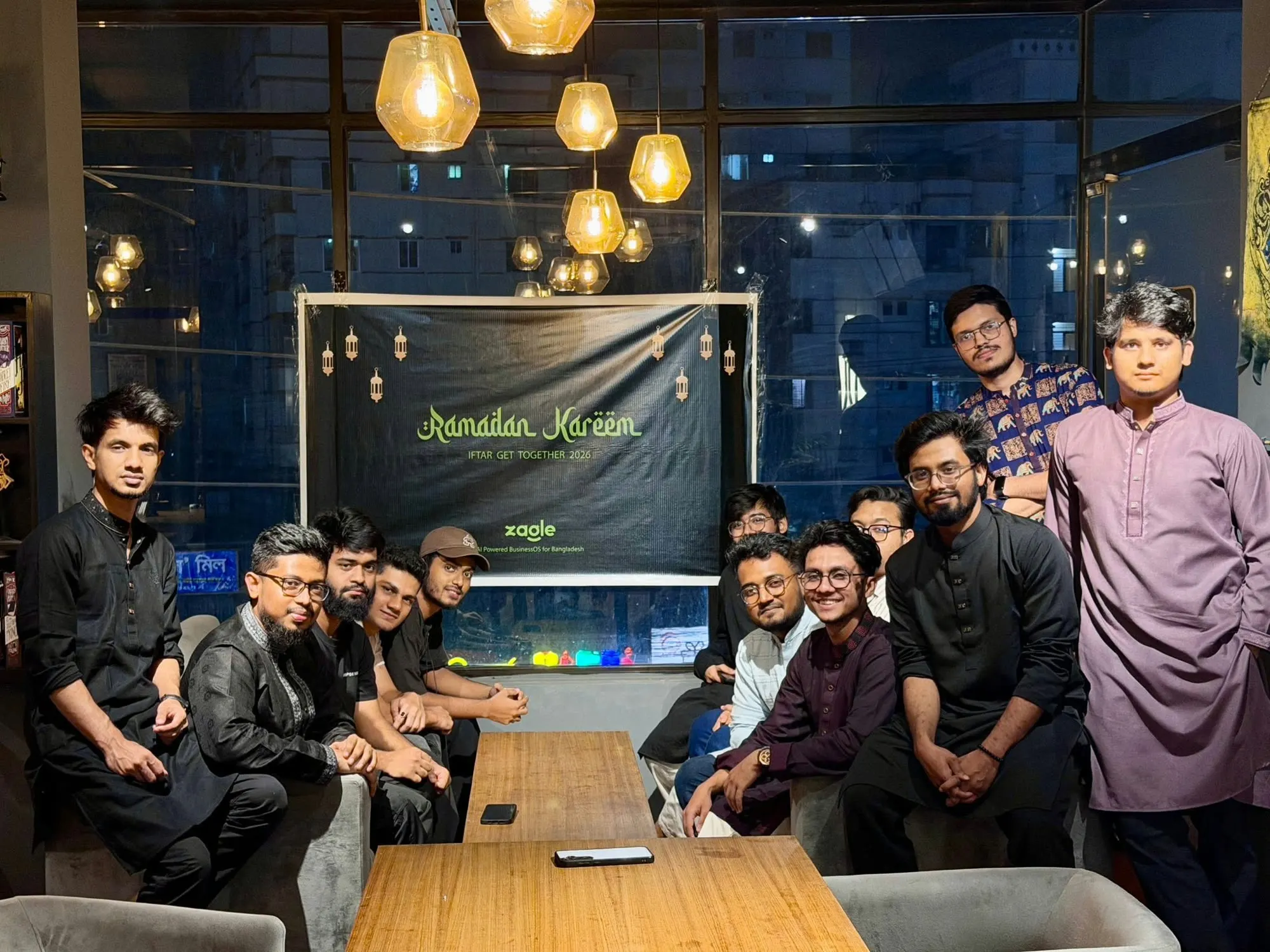 About fifteen people round a cafe table under a banner reading Ramadan Kareem, Iftar get together 2026, Zagle.